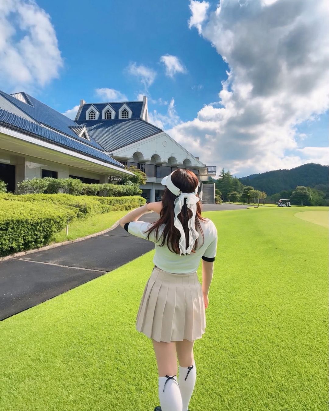 Chamuleのバイカラーリブトップスを着用し、ゴルフ場で後ろを向いている女性。ベージュのプリーツスカートとサンバイザーを合わせたスポーティーなゴルフウェア。