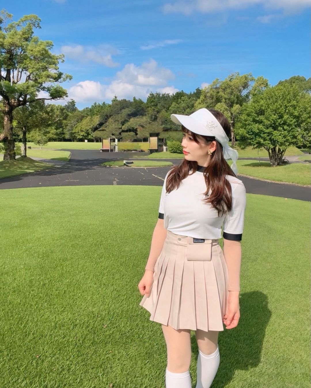 Chamuleのバイカラーリブトップスを着用し、ゴルフ場でポーズをとる女性。白の半袖トップスとベージュのプリーツスカートのゴルフウェア。