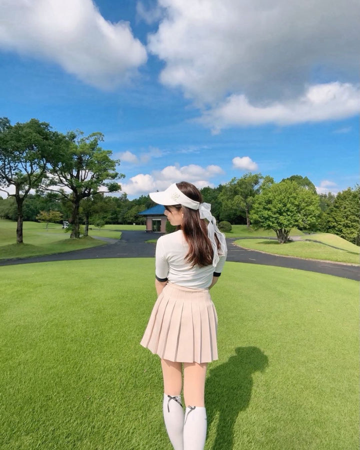 Chamuleのバイカラーリブトップスを着用し、ゴルフ場でポーズをとる女性。白の半袖トップスにベージュのプリーツスカートを合わせたスポーティーなゴルフウェア。
