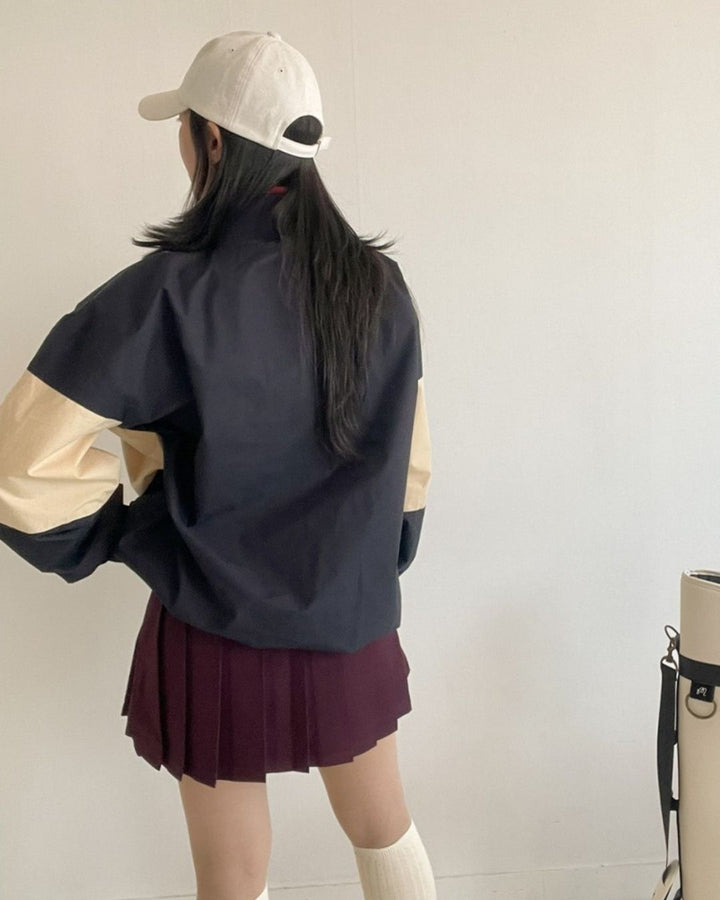 Chamuleの配色切り替えハーフジッププルオーバーを着用したゴルフスタイル。ネイビーとベージュのゆったりとしたシルエットのトップスにバーガンディのプリーツスカートを合わせた後ろ姿。