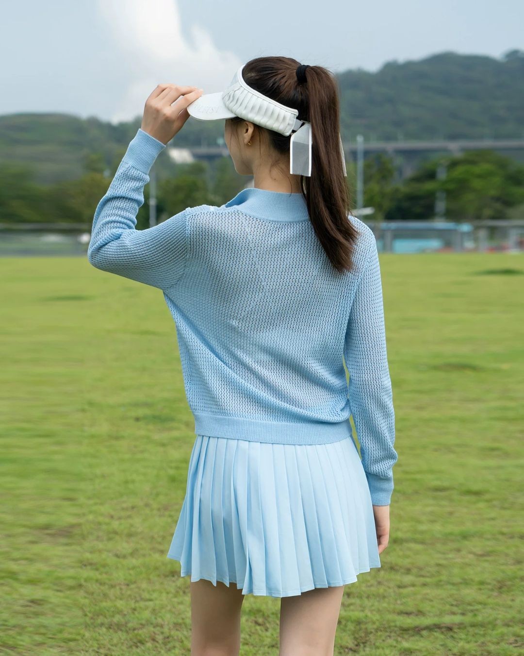 Chamuleのライトブルーのハーフジップニットとプリーツスカートを着用した女性のゴルフウェアの後ろ姿