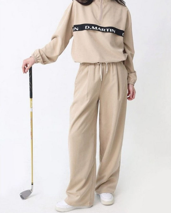 Chamuleのロゴライン入りベージュのハーフジップトップス。ゆったりとしたシルエットで裾にドローコード付き、ゴルフウェアとして着用。