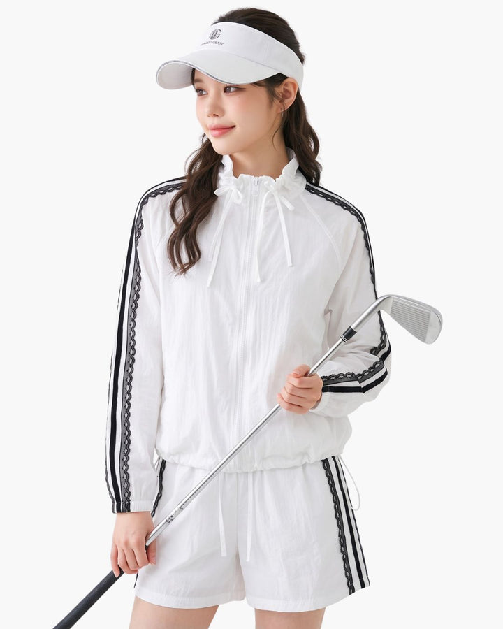 Chamuleの白いレースライン入りジップアップジャケットとショートパンツのゴルフウェアセットアップを着用した女性
